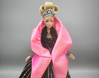 May include: A Barbie doll wearing a black and silver gown with a pink satin shawl. The doll has blonde hair styled in a bun and is wearing a tiara.