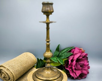 May include: A gold metal candlestick with a round base and a single candle holder. The candlestick is sitting on a piece of burlap fabric.