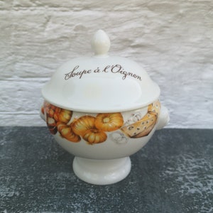 Peut inclure: Soupière en céramique blanche avec un couvercle. La soupière est décorée d'un motif d'oignons et de fromage brun et jaune. Le couvercle porte l'inscription "Soupe à l'Oignon" en cursive brune.