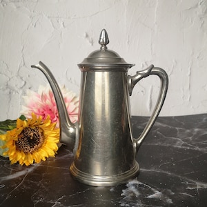 May include: A vintage silver coffee pot with a long spout and a handle. The pot has a lid with a decorative finial. A yellow sunflower and a pink flower are next to the pot on a black marble surface.
