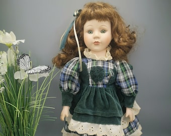 Puede incluir: Una muñeca de porcelana con el pelo largo y rizado marrón, que lleva un vestido de terciopelo verde con una falda a cuadros y un ribete de encaje blanco. Tiene los ojos verdes y lleva zapatos negros.