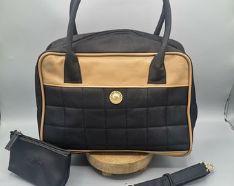 May include: A black quilted handbag with a tan leather trim and a gold logo. The bag has a top handle and a detachable shoulder strap. It comes with a matching black zippered pouch.