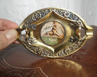 May include: A gold-colored metal ashtray with an oval shape and a raised design of flowers and leaves. The center of the ashtray features a small, round, painted image of a woman reclining on a bed of flowers. The image is surrounded by a gold-colored frame.