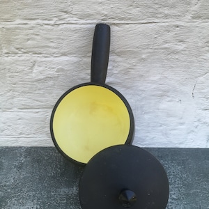 Può includere: Una pentola in ghisa nera con un interno in smalto giallo e un coperchio nero. La pentola ha un manico e una manopola sul coperchio.