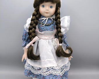 Puede incluir: Muñeca de porcelana con cabello castaño trenzado, ojos azules y un vestido floral azul con un delantal blanco y ribete de encaje. La muñeca lleva un gorro blanco y zapatos blancos. Un collar adorna el cuello de la muñeca.