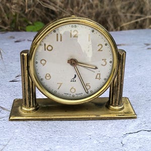 Puede incluir: Un reloj vintage dorado con esfera redonda y números romanos. El reloj tiene una esfera color crema con números y manecillas negras. La base es rectangular y el reloj está marcado "JAZ" y "MADE IN FRANCE".