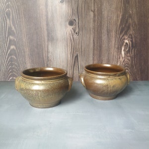Może przedstawiać: Dwie brązowe ceramiczne miski z pstrokatą glazurą. Miski mają po dwie rączki z każdej strony i stoją na szarej powierzchni.