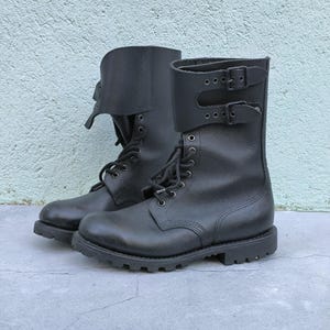 Könnte beinhalten: Ein Paar schwarze Leder-Kampfstiefel mit robustem Design. Die Stiefel haben einen Schnürverschluss, eine dicke Sohle und eine umklappbare Manschette, die mit Schnallen befestigt ist. Vor hellblau-grauem Hintergrund.