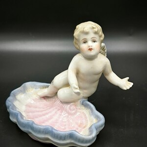 Puede incluir: Una figurita de querubín de porcelana sentada en una concha de mar rosa y azul. El querubín tiene el pelo rubio y lleva un vestido blanco.
