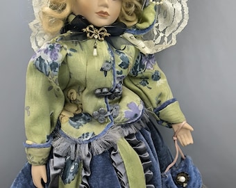 Puede incluir: Una muñeca de porcelana con cabello rubio rizado, ojos azules y un rostro delicado. La muñeca viste un vestido floral verde y azul con un gorro a juego y un pequeño bolso azul. El vestido tiene detalles y adornos intrincados.