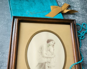 May include: A framed oval portrait in a dark wood frame. The portrait is of a woman in a draped garment. A teal book with a gold bow and a strand of turquoise beads are also in the image. The book has the word "Memories" on it.
