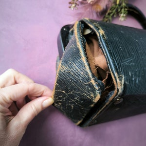 May include: A black leather vintage camera case with a worn and distressed look. The case is open and the inside is lined with brown fabric.