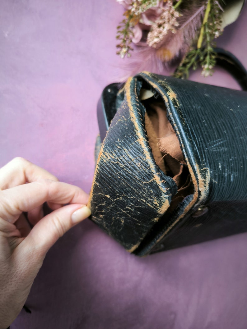 May include: A black leather vintage camera case with a worn and distressed look. The case is open and the inside is lined with brown fabric.