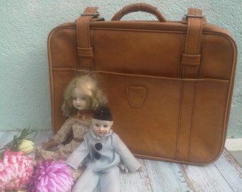 Może przedstawiać: Brązowa skórzana walizka w stylu vintage o klasycznym designie, stojąca na jasnoniebieskiej drewnianej powierzchni. Dwie porcelanowe lalki, jedna z blond włosami i białą sukienką, a druga w niebiesko-białym stroju klauna, siedzą przed walizką.