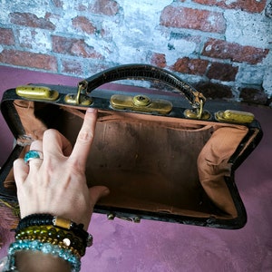 May include: A vintage black leather doctor's bag with brass hardware and a brown fabric interior. The bag is open and empty.