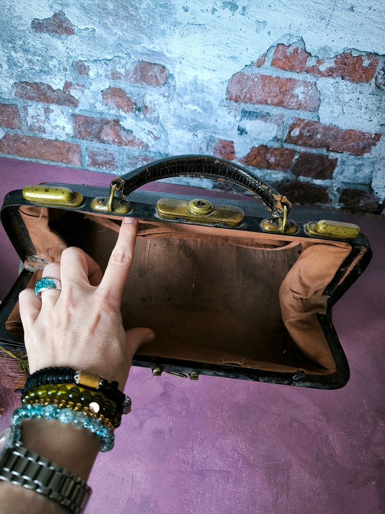 May include: A vintage black leather doctor's bag with brass hardware and a brown fabric interior. The bag is open and empty.