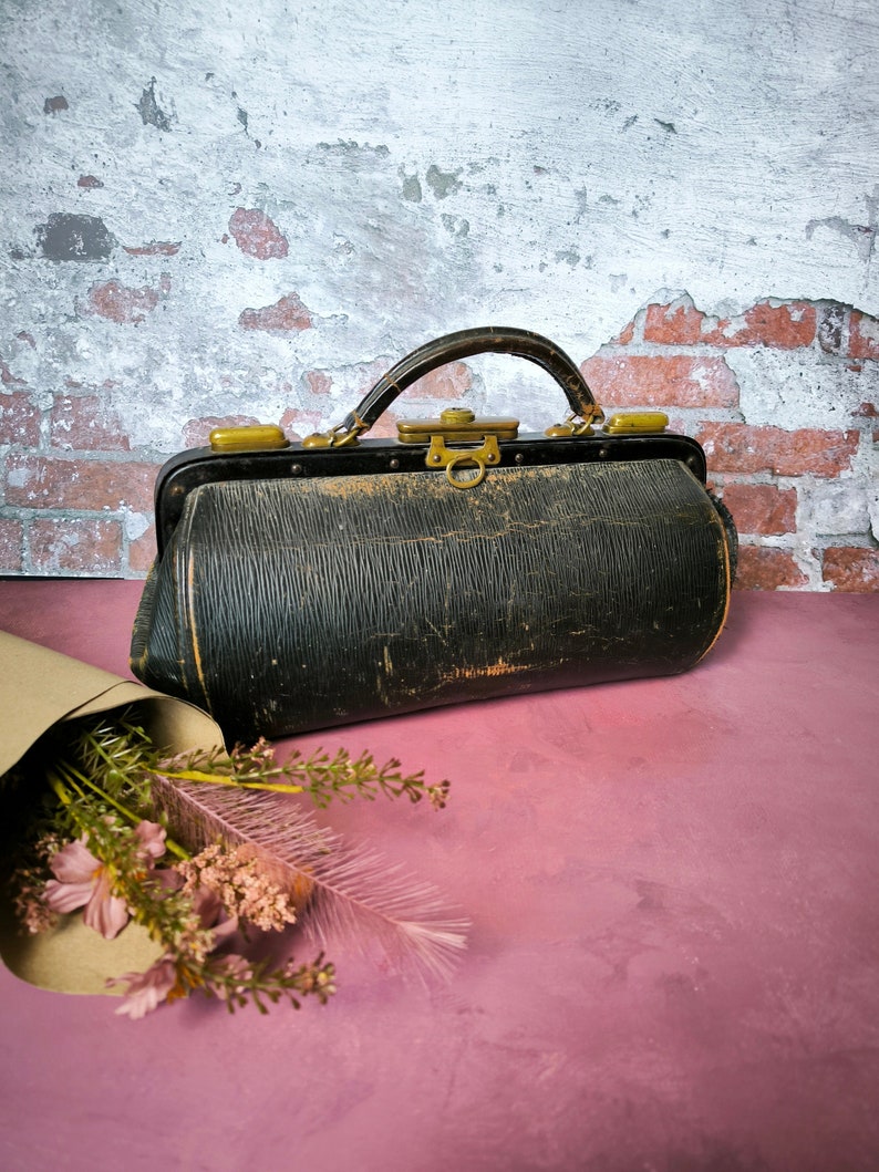 May include: A vintage black leather doctor's bag with brass hardware and a worn, distressed look. The bag is sitting on a pink surface with a bouquet of pink flowers in the foreground.