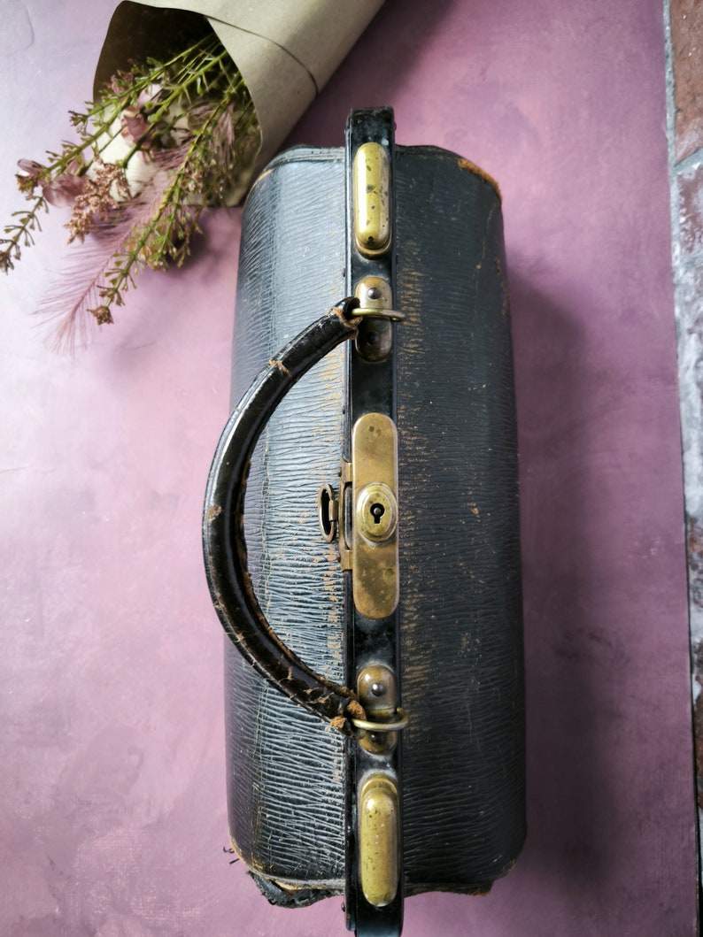 May include: A vintage black leather doctor's bag with brass hardware and a handle. The bag has a worn and aged look, with visible scratches and scuffs.