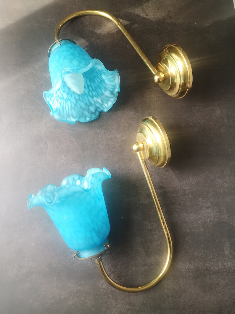 May include: Two wall sconces featuring brass fixtures and turquoise glass shades. The shades are flower-shaped with a speckled pattern. The sconces have a curved arm design and are mounted on round brass bases.