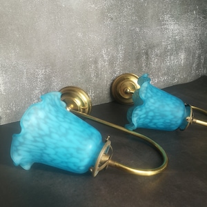 May include: Two vintage wall sconces with a brass finish and turquoise glass shades. The shades have a speckled pattern and a ruffled edge. The sconces are mounted on a dark surface against a textured grey wall.