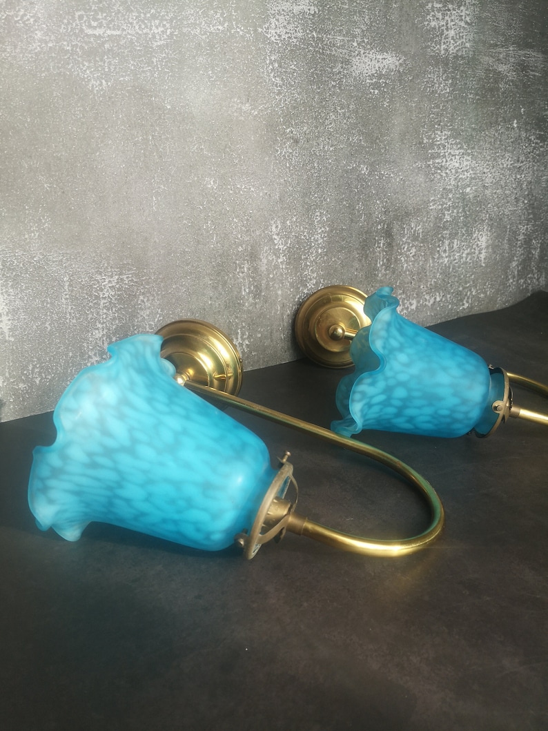 May include: Two vintage wall sconces with a brass finish and turquoise glass shades. The shades have a speckled pattern and a ruffled edge. The sconces are mounted on a dark surface against a textured grey wall.