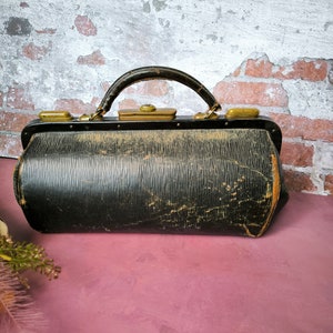 May include: A vintage black leather doctor's bag with brass hardware and a worn, distressed look. The bag has a top handle and a rectangular shape.