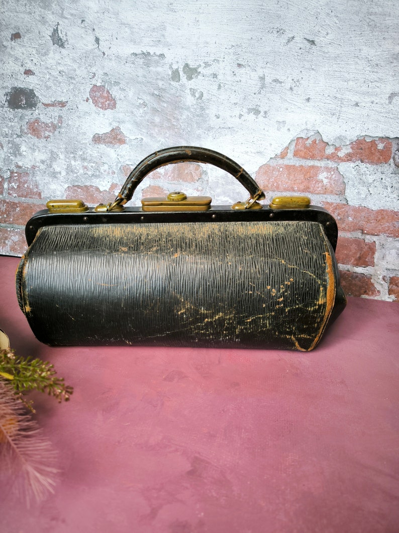 May include: A vintage black leather doctor's bag with brass hardware and a worn, distressed look. The bag has a top handle and a rectangular shape.