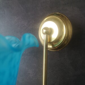 May include: A vintage wall sconce with a brass finish and a light blue glass shade. The sconce features a round backplate, a slender arm, and a bell-shaped shade. The glass shade has a ruffled edge.