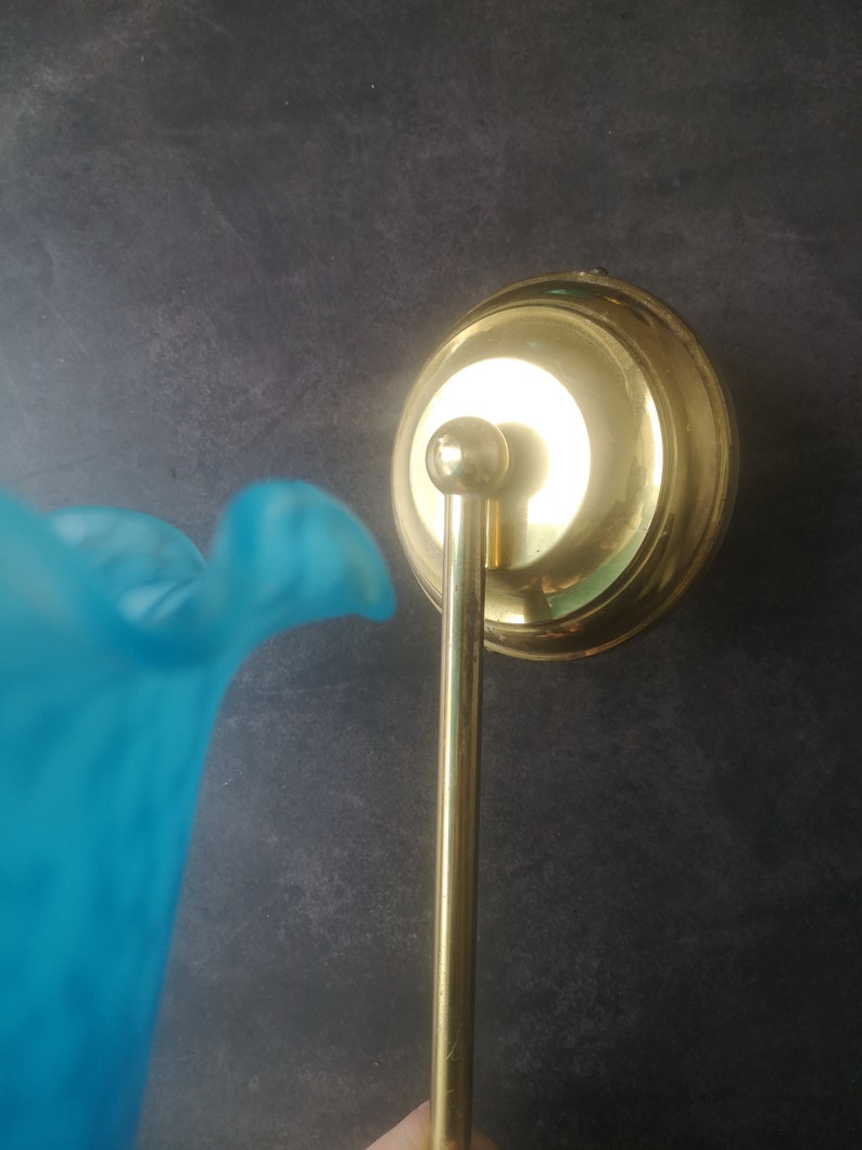 May include: A vintage wall sconce with a brass finish and a light blue glass shade. The sconce features a round backplate, a slender arm, and a bell-shaped shade. The glass shade has a ruffled edge.