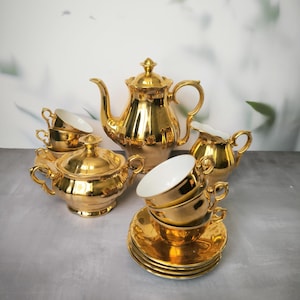 Puede incluir: Un juego de té chapado en oro con tetera, jarra de crema, azucarero y dos tazas y platillos. El juego está hecho de porcelana y tiene un acabado brillante y metálico.