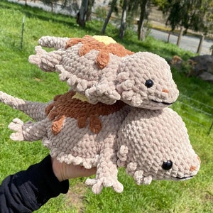 Pode incluir: Dois lagartos de pelúcia de crochê, um em cima do outro. O lagarto de cima é marrom e bege com uma mancha amarela nas costas. O lagarto de baixo é bege com manchas marrons. Ambos os lagartos têm olhos pretos.