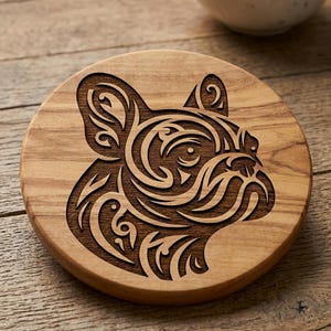 Peut inclure: Un dessous de verre rond en bois présentant un motif gravé au laser représentant une tête de bouledogue français. L'illustration du chien est composée de lignes et de motifs fluides de style tribal. Le grain du bois est visible.