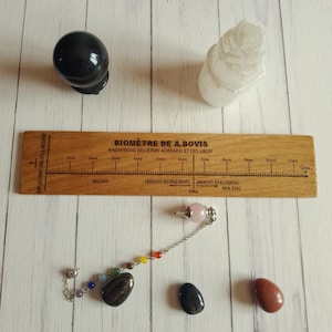 Peut inclure: Une sphère noire, une tour de cristal blanc translucide, une règle en bois échelle de Bovis, un pendule avec une pierre rose et des perles multicolores, et trois pierres lisses noire, gris foncé et brun rougeâtre sur une surface blanche.