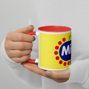 Könnte beinhalten: Eine weiße Keramiktasse mit rotem Rand und Henkel. Die Tasse hat einen gelben Hintergrund mit einem roten und blauen Logo, auf dem "I  <3  Milky" steht.