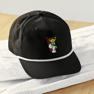 Può includere: Cappellino da golf nero con dettaglio in corda bianca e visiera curva. Il cappellino presenta un disegno ricamato di un orso giallo che indossa un abito da golf e porta una sacca da golf. Il cappellino è appoggiato su una superficie bianca.