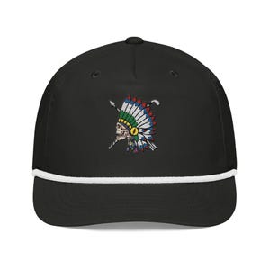 May include: A black baseball cap featuring a white rope detail across the base of the crown. The front displays an embroidered graphic of a human skull wearing a colourful feathered headdress, positioned in front of two crossed golf clubs.