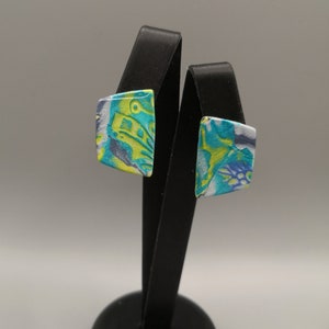 May include: A pair of square-shaped earrings with a blue, green and white abstract pattern. The earrings are displayed on a black stand.