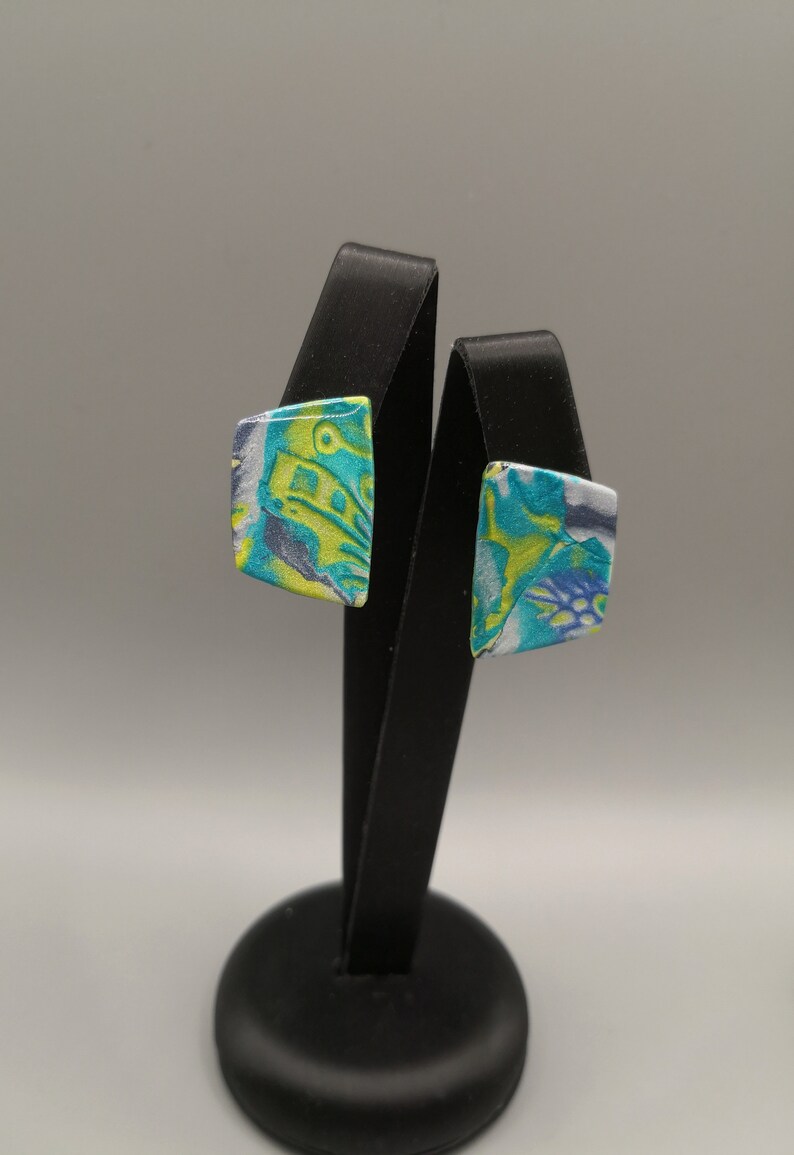 May include: A pair of square-shaped earrings with a blue, green and white abstract pattern. The earrings are displayed on a black stand.
