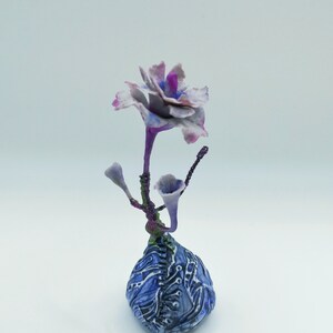 Könnte beinhalten: Eine Keramik-Blumenskulptur mit einem blauen und weißen gemusterten Sockel. Die Blume ist lila und weiß mit einem grünen Stiel.