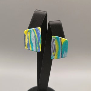May include: A pair of square-shaped earrings with a marbled design in shades of blue, green, and yellow. The earrings are displayed on a black stand.