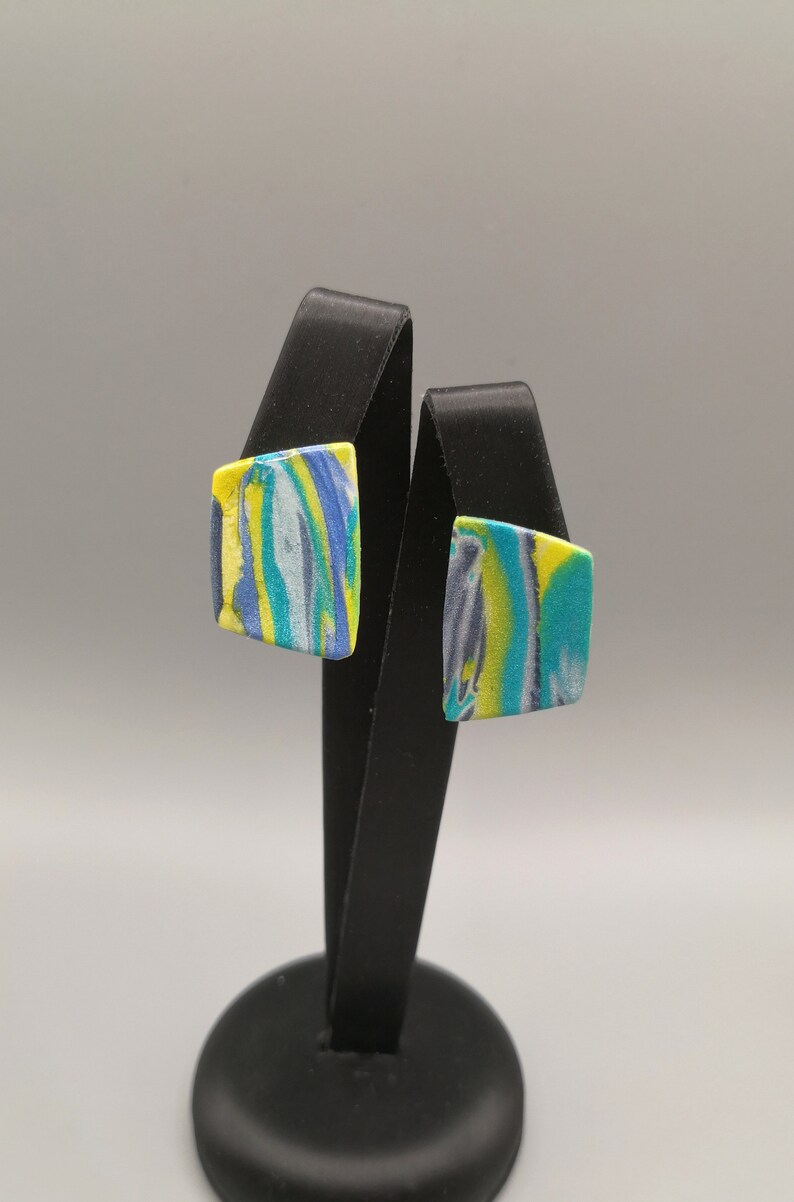 May include: A pair of square-shaped earrings with a marbled design in shades of blue, green, and yellow. The earrings are displayed on a black stand.
