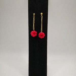 May include: A pair of gold chain earrings with red flower-shaped charms. The earrings are displayed on a black velvet stand.