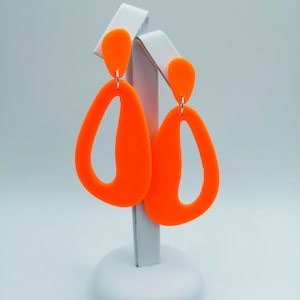 Può includere: Un paio di orecchini geometrici di colore arancione brillante con un design a taglio. Gli orecchini sono esposti su un supporto bianco.