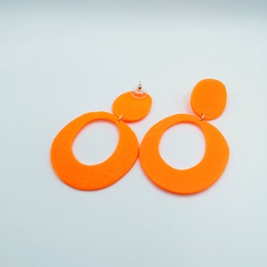 May include: A pair of neon orange dangle earrings with a large oval shape and a smaller circle shape. The earrings are made of a lightweight material and have a modern, geometric design.