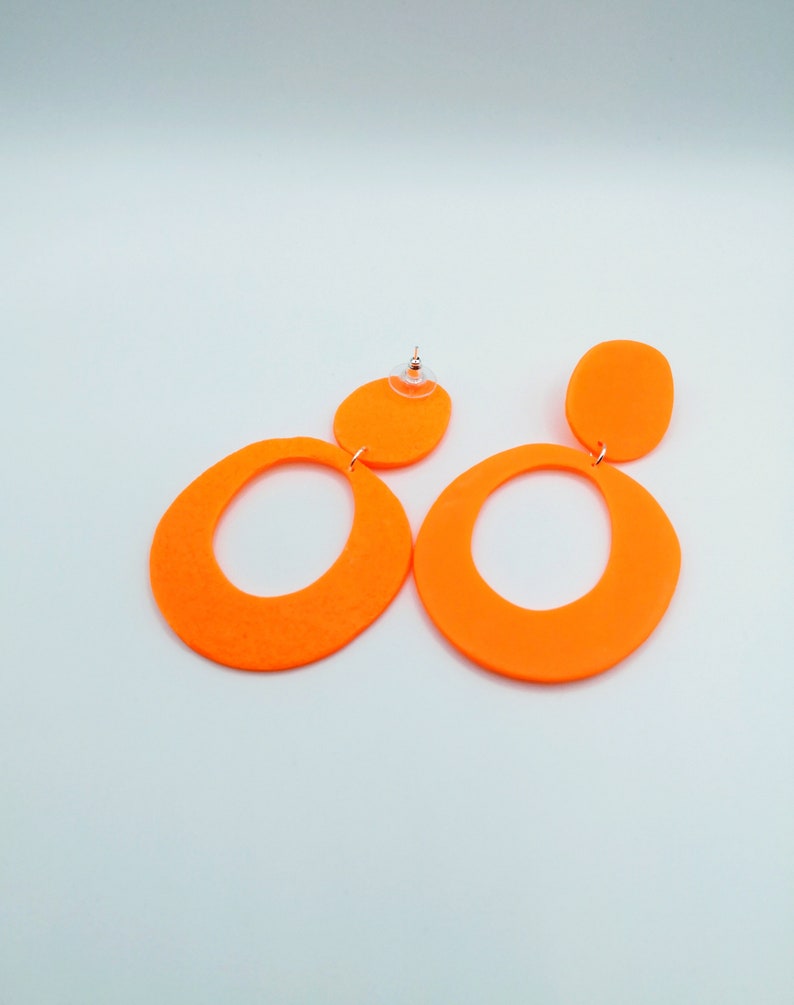 May include: A pair of neon orange dangle earrings with a large oval shape and a smaller circle shape. The earrings are made of a lightweight material and have a modern, geometric design.