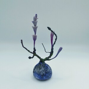 Könnte beinhalten: Eine abstrakte Skulptur mit einer gemusterten, blauen und grauen, kugelförmigen Basis. Dunkle, astähnliche Strukturen erheben sich von der Basis und tragen zarte, längliche, violette Formen, die eine markante, organische Ästhetik erzeugen.