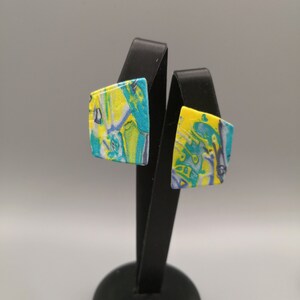 May include: A pair of square earrings with a marbled pattern in yellow, blue and white. The earrings are displayed on a black stand.