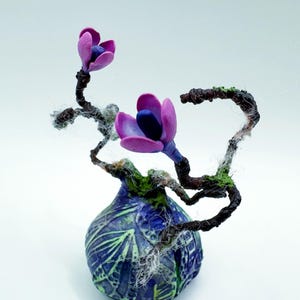Könnte beinhalten: Eine kleine, blaue und grüne Keramikvase mit einem wirbelnden Muster. Die Vase hält einen zarten Zweig mit zwei rosa Blüten und ein paar zarten Ranken.