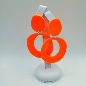 May include: A pair of bright orange statement earrings with a large circle and a smaller oval shape. The earrings are displayed on a white stand.