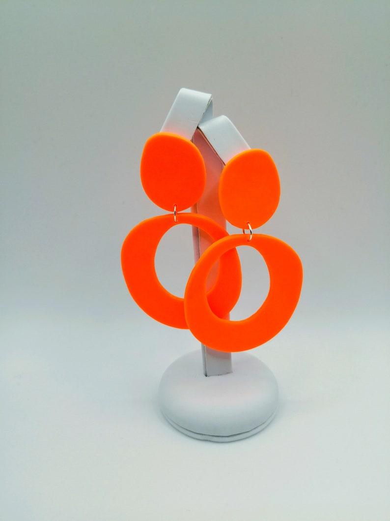 May include: A pair of bright orange statement earrings with a large circle and a smaller oval shape. The earrings are displayed on a white stand.